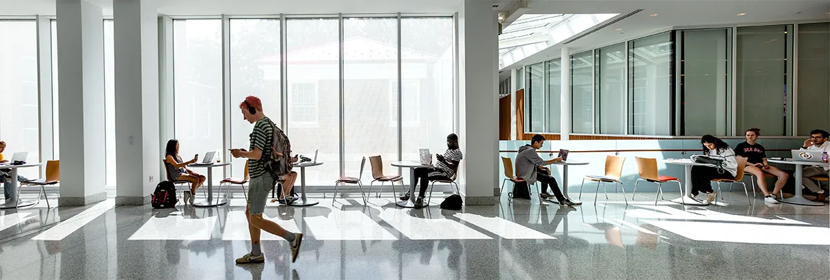 Students sitting at tables in front of large windows. One student is walking across