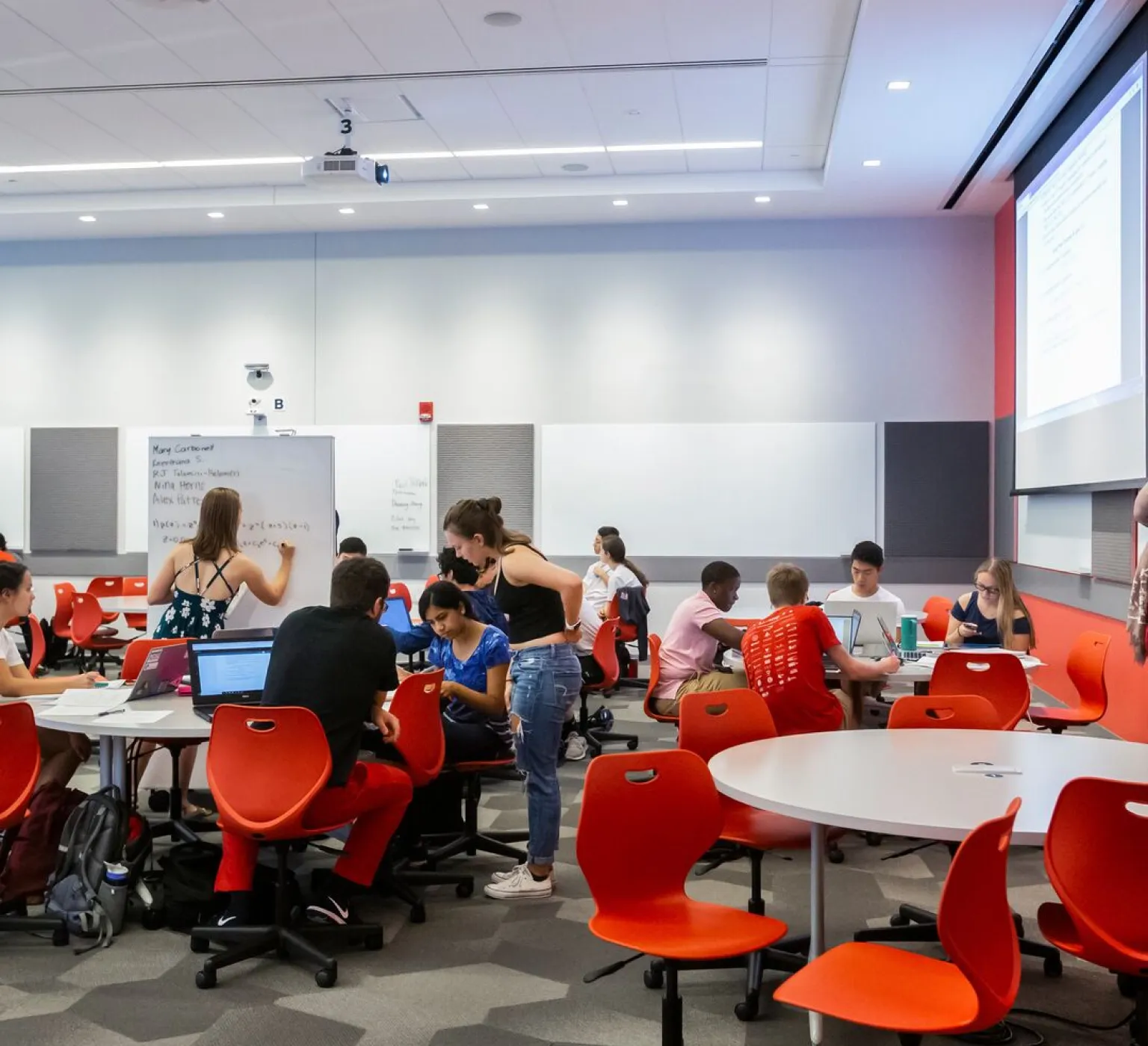 Students active in group work in a classroom with white boards. Some are seated working together. One at a whiteboard